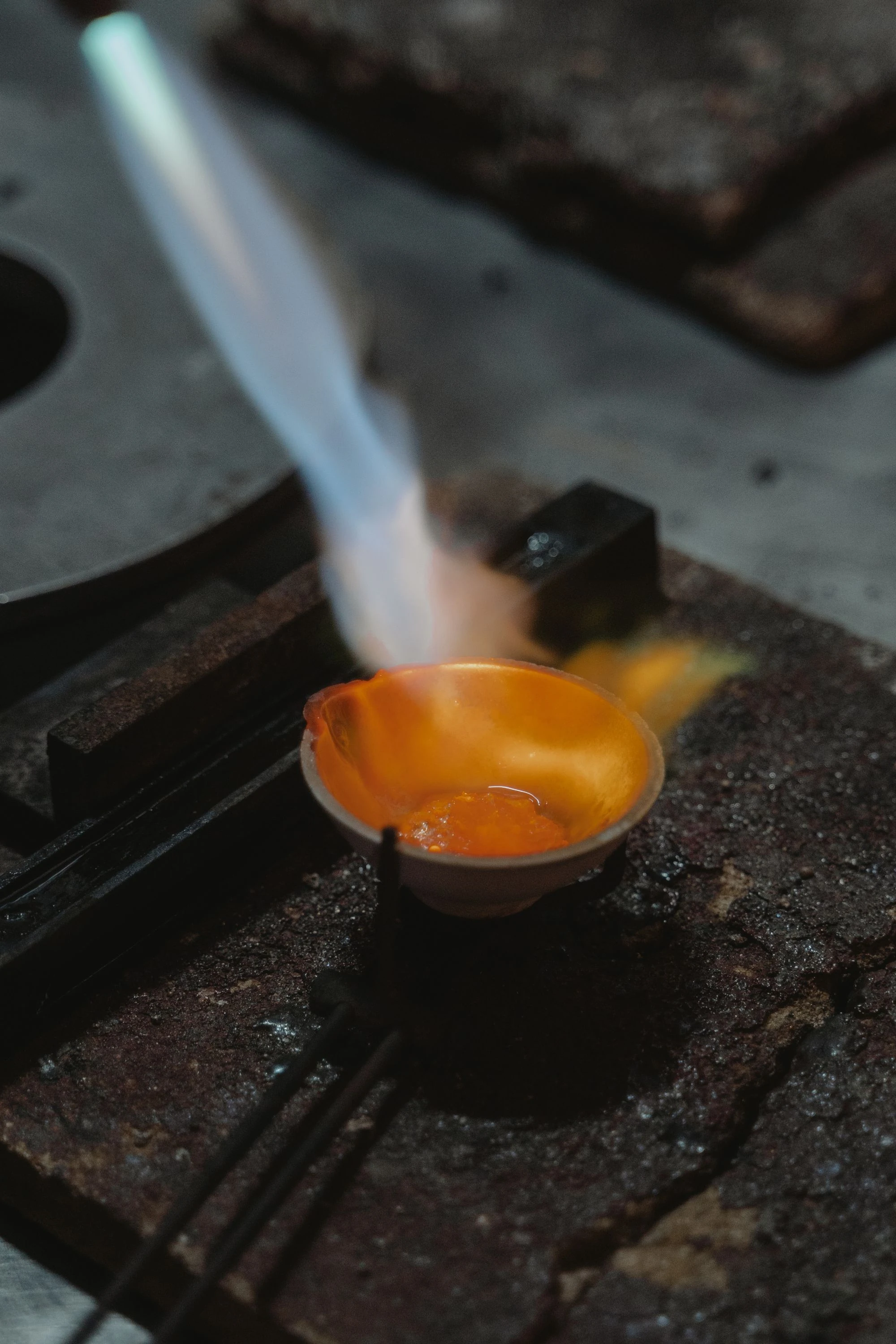 Torch flame heating metal in a small ceramic melting dish at a jewelry workbench